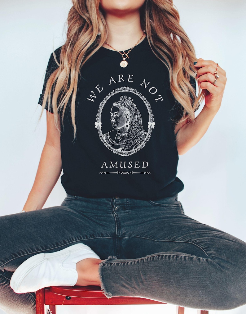May include: A black t-shirt with a white graphic of a queen's portrait in a frame with the text "We Are Not Amused".