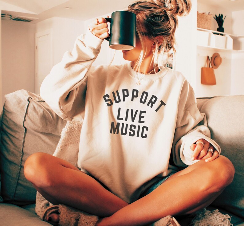 May include: A woman wearing a light beige sweatshirt with the text "SUPPORT LIVE MUSIC" printed on the front. She is sitting on a couch and holding a black mug.
