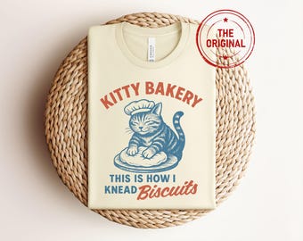 May include: Cream-colored t-shirt with the text "KITTY BAKERY" and a blue and white illustration of a cat wearing a chef's hat. Below the image, the text reads "THIS IS HOW I KNEAD BISCUITS". A red stamp says "THE ORIGINAL".