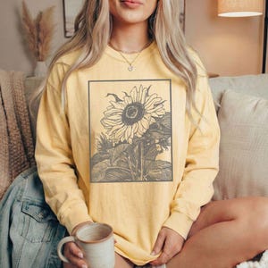May include: A long-sleeve crewneck sweatshirt in a pale yellow color featuring a rectangular graphic print on the chest. The print displays a detailed, dark gray line-art illustration of a large sunflower with leaves and a stem.