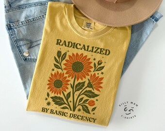 May include: Mustard yellow t-shirt with the text "RADICALIZED BY BASIC DECENCY" and a floral design featuring sunflowers. The shirt is folded on top of a pair of blue jeans and a tan hat.
