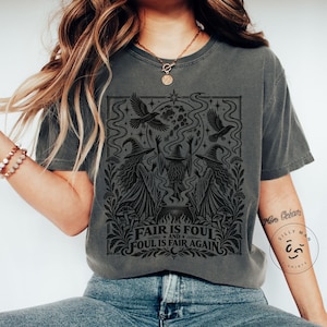 May include: A charcoal grey short-sleeve t-shirt featuring a black graphic print of three figures in pointed hats standing around a cauldron. The design includes ravens, a moon, mushrooms, and the text Fair is foul and foul is fair again.