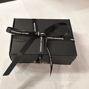 Op de afbeelding: Een zwarte geschenkdoos vastgebonden met een zwart lint. Op het lint staat "Enrichment Candles". Een kleine gouden sticker met de woorden "Thank You" staat op de doos.