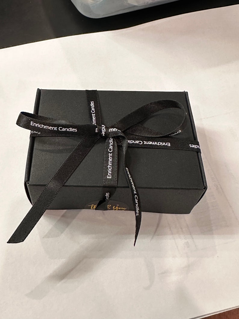 Op de afbeelding: Een zwarte geschenkdoos vastgebonden met een zwart lint. Op het lint staat "Enrichment Candles". Een kleine gouden sticker met de woorden "Thank You" staat op de doos.