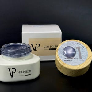 May include: A collection of Vibe Polish nail products. The image shows a white jar with a dark blue polish, a white box, and a wooden jar with a silver-blue polish. The text on the wooden jar reads "Glass Bead Cat Eye Collection".