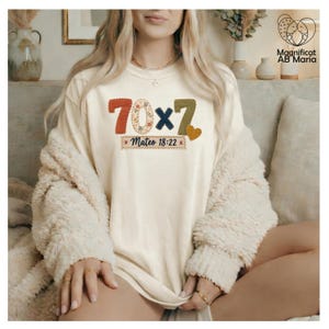 Puede incluir: Una camiseta color crema con un gráfico que muestra el texto 70x7 y Mateo 18:22. Los números tienen una textura de costura en naranja, estampado floral, azul marino y verde oliva, junto a un pequeño icono de corazón naranja.