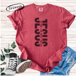 Puede incluir: Camiseta de color rojo carmesí con la palabra "JESUS" impresa verticalmente en marrón oscuro, acompañada del texto "THE WAY - THE TRUTH - THE LIFE". La camiseta se combina con pantalones cortos de mezclilla y zapatillas deportivas blancas y negras.