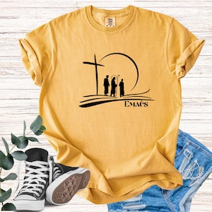 Puede incluir: Camiseta de color amarillo mostaza con un gráfico negro que representa una cruz, figuras y un sol. La palabra "EMAÚS" está impresa debajo. También se ven zapatillas negras y blancas y un pantalón corto vaquero.