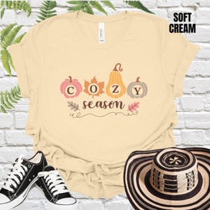 Puede incluir: Camiseta color crema con las palabras "COZY season" y gráficos de calabazas y hojas. La camiseta se combina con zapatillas negras y blancas y un sombrero a rayas. Las palabras "SOFT CREAM" están impresas en un recuadro negro.