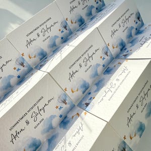 May include: A collection of white rectangular cards featuring a watercolor design of blue clouds, a hot air balloon, stars, and a crescent moon. The text reads SÜNNETİMİZE HOŞGELDİNİZ, Adem & Süleyman, and the date 02.04.2026 in black script and print.