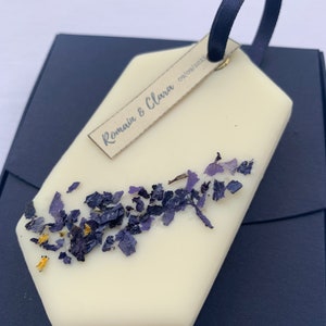 Peut inclure: Un objet de forme géométrique de couleur crème, avec un ruban bleu foncé et une petite étiquette indiquant "Romain & Clara 09/09/2023". L'objet est décoré de pétales de fleurs séchées violettes et jaunes. Il repose sur une surface bleu foncé.