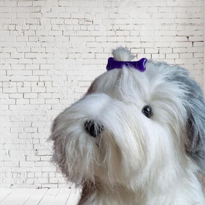 Op de afbeelding: Een pluche hondenspeeltje met lang, ruig wit en grijs bont. De hond heeft een zwarte neus en donkere ogen, met een kleine, glanzende paarse botvormige haarclip bevestigd aan het haar boven op de kop, wat zorgt voor een opvallend uiterlijk.