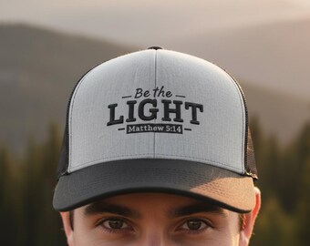 May include: A gray and black trucker hat with the text "Be the LIGHT" and "Matthew 5:14" in black. The hat has a mesh back and a curved brim.
