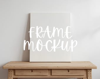 Könnte beinhalten: Eine weiße Leinwand mit dem Text "FRAME MOCKUP" in Schreibschrift, die auf einem hellbraunen Holztisch mit zwei Schubladen steht. Der Hintergrund ist eine schlichte weiße Wand, die eine saubere, minimalistische Ästhetik erzeugt.