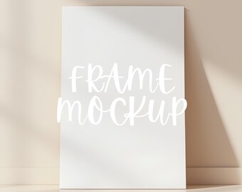 May include: White frame mockup on a light beige background. The frame is standing upright and has a shadow cast on the wall behind it. The text "FRAME MOCKUP" is written in white on the frame.