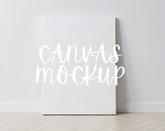 Könnte beinhalten: Ein weißes Leinwand-Mockup, das an einer weißen Wand lehnt. Der Text "CANVAS MOCKUP" ist in weißer Schrift auf der Leinwand geschrieben. Der Boden ist helles Holz mit Fischgrätmuster.