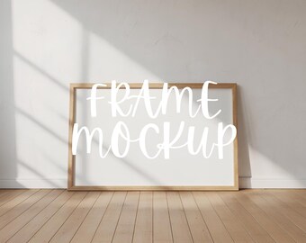 May include: A wooden frame mockup with the words "FRAME MOCKUP" in white script. The frame is against a white wall, with a wooden floor. The image has natural light and shadows. The frame is rectangular.