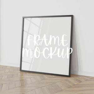 May include: A black framed square mockup leaning against a white wall on a wooden floor. The words "FRAME mockup" are written in white script across the center of the frame.