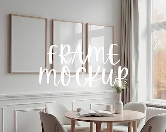 May include: Three empty picture frames in a living room setting. The frames are rectangular and have a light wood finish. The frames are hanging on a white wall above a wooden table with four chairs. The text "FRAME MOCKUP" is written in white cursive letters on the wall.