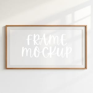 Puede incluir: Un marco rectangular de madera con interior blanco, que muestra el texto "FRAME MOCKUP" en un estilo manuscrito. El marco está sobre una pared blanca, con luz natural proyectando una sombra.
