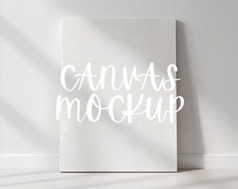 Könnte beinhalten: Ein weißes Leinwand-Mockup, das an einer weißen Wand lehnt. Die Leinwand hat eine strukturierte Oberfläche und die Worte "CANVAS MOCKUP" sind in einer Schreibschrift auf die Vorderseite geschrieben. Schatten von einem Fenster fallen auf die Wand und die Leinwand.