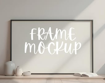 May include: A black frame mockup with the text "FRAME MOCKUP" in white lettering on a white background. The frame is sitting on a wooden floor with a book and two vases in front of it.
