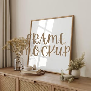 May include: A wooden framed art print mockup with the words "FRAME MOCKUP" in a brown script font. The frame is leaning against a light-colored wall, with decorative items on a wooden cabinet in front of it, including vases and books.