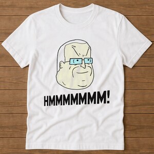 May include: White t-shirt with a cartoon image of a man wearing glasses and the word "HMMMMMMMM!" in black. The design is printed on the front. The shirt has short sleeves and a round neck.
