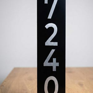 May include: Black rectangular address sign with silver numbers 7240. The sign is standing upright on a wooden surface. The numbers are large and easy to read.