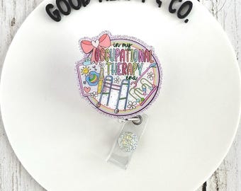 Puede incluir: Un portatarjetas colorido con el texto "In my Occupational Therapy era". El diseño incluye un lazo rosa, un arcoíris e ilustraciones de herramientas de terapia. El portatarjetas tiene una correa de plástico transparente y un botón decorativo plateado brillante.