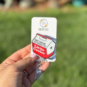 May include: A white and red badge reel with a glitter finish. The badge reel features a cartoon image of a cooler with the text "Don't hate me just because I'm a Little Cooler".