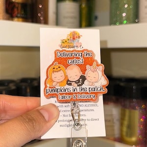 Puede incluir: Un portatarjetas con un diseño de calabaza naranja brillante. La tarjeta dice "Delivering the cutest pumpkins in the patch. Labor & Delivery" con ilustraciones de bebés. El carrete tiene un clip de plástico transparente.