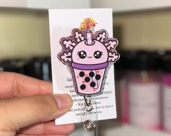 May include: A cute, pink and purple acrylic badge reel featuring a cartoon boba tea design. The badge reel has a glittery border and a smiling face on the boba cup. The reel is attached to a white card with text.