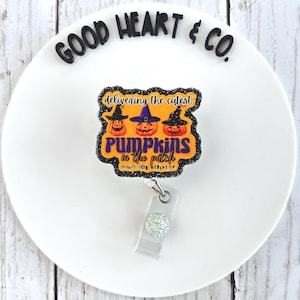 Puede incluir: Un portatarjetas con temática de Halloween con el texto "Delivering the cutest pumpkins in the patch". El diseño presenta tres calabazas naranjas con sombreros de bruja. El carrete tiene una base de plástico transparente y un acento brillante.