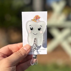 May include: A white, glittery tooth-shaped badge reel with a smiling face and pink cheeks. A small bouquet of flowers sits atop the tooth. The badge reel has a clear plastic retractable cord and a metal clip.