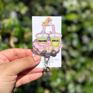 May include: A clear acrylic badge reel featuring two matcha drinks with straws, the words "Matcha" and "Girlie" are printed on the cups. The badge reel has a pink glitter border and a floral design. The reel is attached to a white card.
