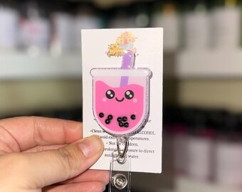 May include: A pink bubble tea badge reel with a smiling face and black tapioca pearls. The badge reel has a clear plastic clip and a retractable cord. The design is on a white card with text.