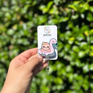 Puede incluir: Un carrete de insignia de brillo rosa y verde con un perezoso que lleva un flotador de flamenco rosa. El carrete tiene el texto "GOOD HEART" y "BADGE REEL" en él. El carrete está unido a un porta insignias de plástico transparente.