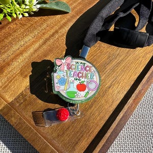 May include: A round, glittery badge reel with the text "in my SCIENCE TEACHER era" and science-themed illustrations. The badge reel is attached to a black lanyard and a clear plastic clip with a red, textured accent.