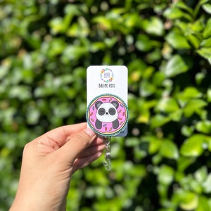 May include: A pink and green donut with sprinkles and a panda bear inside. The donut is attached to a silver retractable badge reel with the text "GOOD HEARTS BADGE REEL".