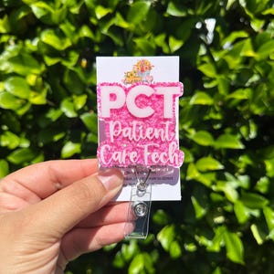 Puede incluir: Un portatarjetas rosa y blanco con el texto "PCT Patient Care Tech". El portatarjetas tiene una correa de plástico transparente y un clip metálico. El fondo es verde borroso.