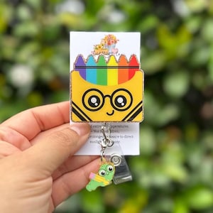 May include: A yellow badge reel with a crayon box design, featuring a rainbow of colors and a smiling face with glasses. A small green worm charm hangs below. The badge reel is held against a blurred green background.