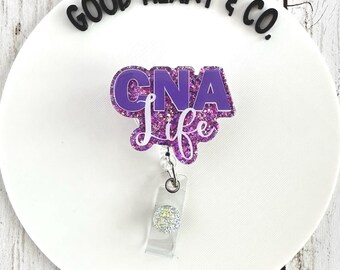 May include: A purple and white CNA Life badge reel with glitter accents. The badge reel has a clear plastic clip and a decorative, sparkly button. The words "Good Heart & Co." are above the badge reel.