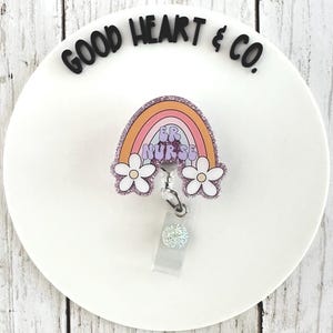 May include: A badge reel with a rainbow design and the text "ER Nurse". The rainbow has orange, pink, and purple colors with glitter accents. Two white daisy flowers are on the bottom of the badge reel.