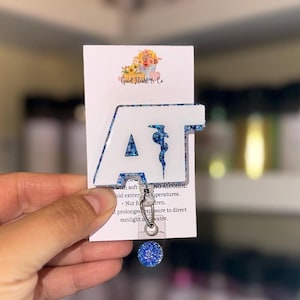 May include: A white badge reel with the letters "AT" in a blue floral pattern. The badge reel has a retractable cord and a blue glitter button. The card behind the badge reel has the text "Good Heart & Co."