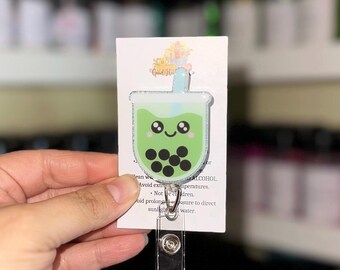 May include: A green and white badge reel featuring a cute boba tea design with black tapioca pearls. The badge reel has a silver clip and is attached to a white card with text. The text includes warnings about alcohol, extreme temperatures, and sunlight.