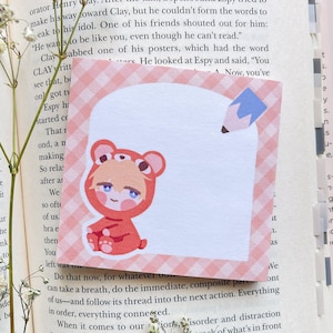 Puede incluir: Una nota adhesiva cuadrada con un borde rosa a cuadros y una ilustración de dibujos animados de una persona con un disfraz de oso. Un lápiz azul está en la parte superior derecha. La nota está en un libro abierto.