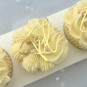 May include: Three white cupcakes with gold frosting and gold glitter. The cupcakes are decorated with gold foil letters that spell "Girl".