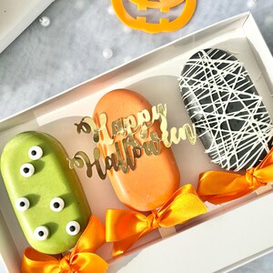 May include: Three Halloween-themed popsicles in a white box. The popsicles are decorated with green eyes, black and white spiderwebs, and orange with the words "Happy Halloween".
