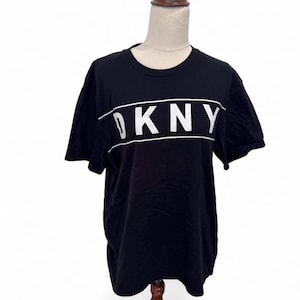 May include: Black t-shirt featuring the white text "DKNY" across the chest. The text is in a bold, sans-serif font and is centered. The shirt has short sleeves and a crew neck.